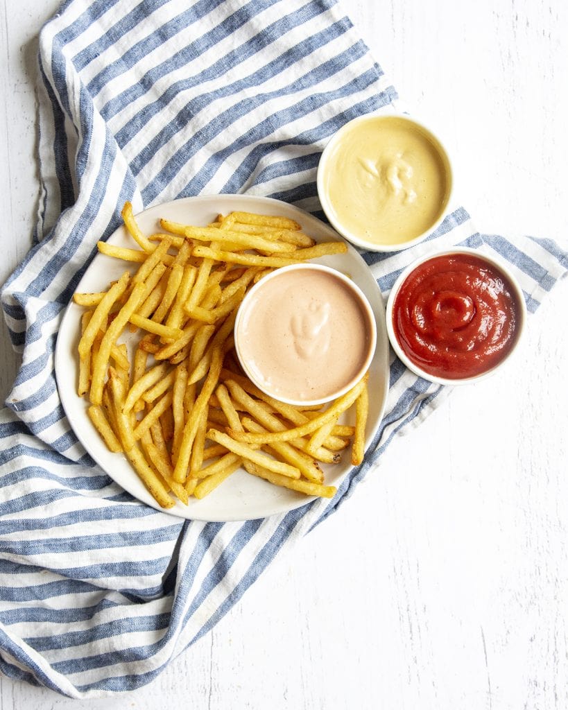 An overhead photo of french fries on a plate, next to a bowl of french fry dipping sauce, aioli, and ketchup.