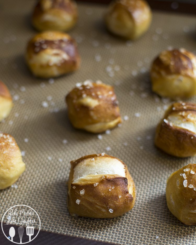 Small soft pretzel bites topped with big pieces of salt, all on a baking tray.