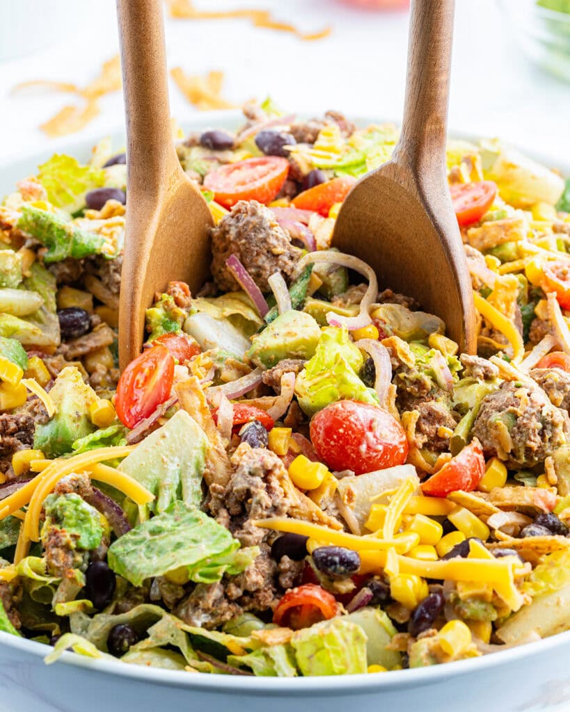 A bowl of tossed beef taco salad with two wooden spoons in it.