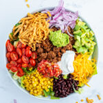 Beef taco salad toppings all in portions on top of a bowl of lettuce, like tomatoes, lettuce, red onion, avocado, cheese, and tortilla s trips.