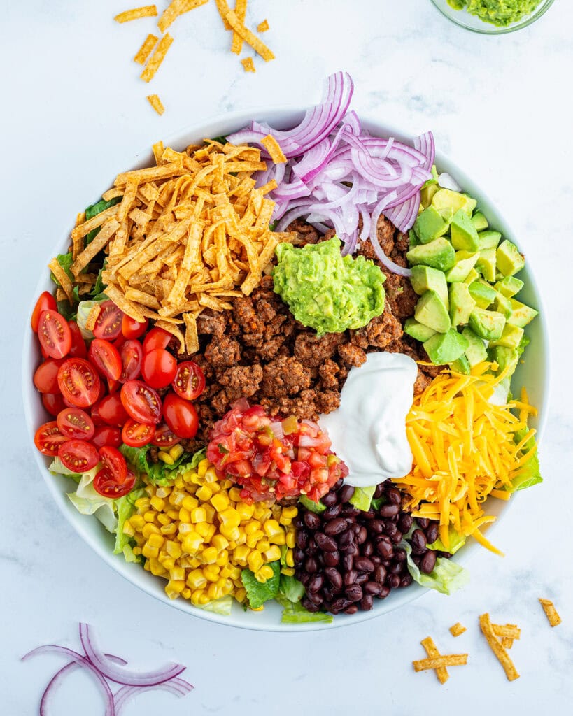 Beef taco salad toppings all in portions on top of a bowl of lettuce, like tomatoes, lettuce, red onion, avocado, cheese, and tortilla s trips. 