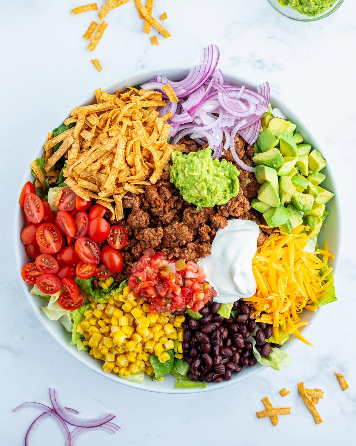 Beef taco salad toppings all in portions on top of a bowl of lettuce, like tomatoes, lettuce, red onion, avocado, cheese, and tortilla s trips.