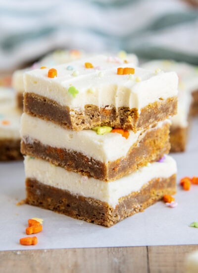 A stack of three carrot cake cookie bars topped with cream cheese frosting, and the top has a bite out of it.