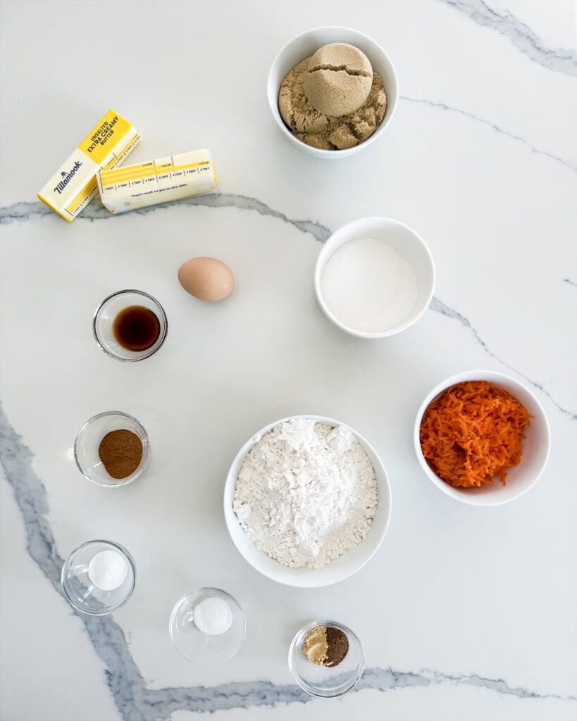 Small bowls of the ingredients needed to make carrot cake cookie bars; like brown sugar, butter, one egg, carrots, flour, cinnamon, and vanilla extract.