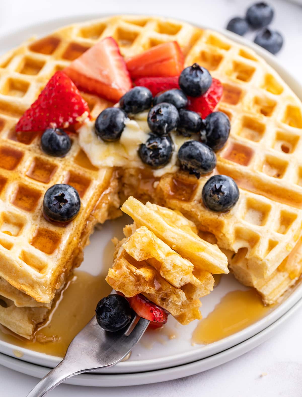 A plate of butter waffles topped with berries, and a bite of waffles on a fork next to it.