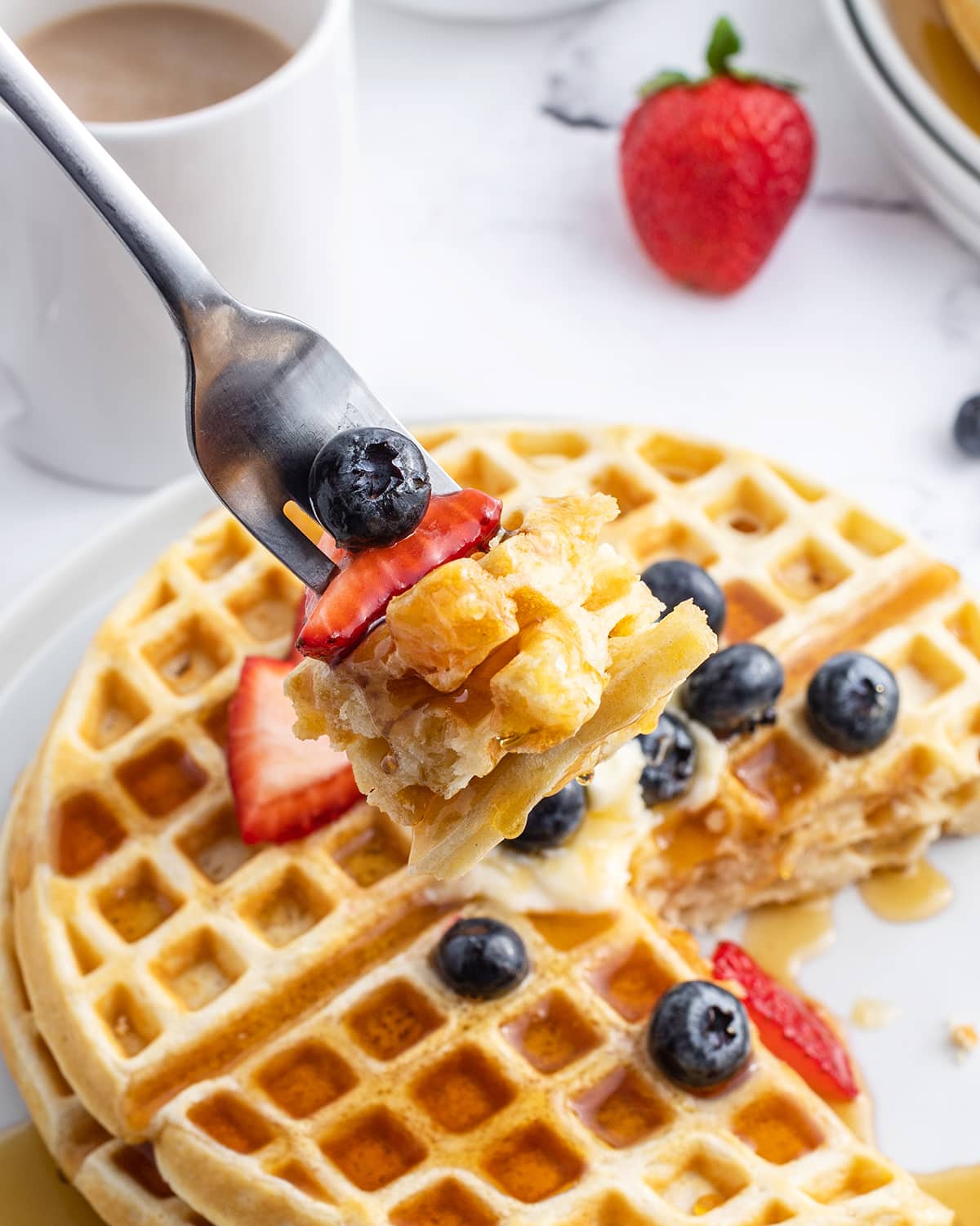 A bite of waffle pieces and fresh berries on a fork above a plate of waffles.