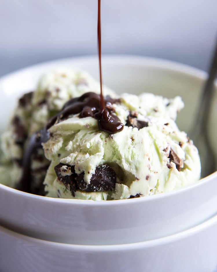 A close up of mint chocolate chip ice cream in a white bowl with chocolate hot fudge being drizzled on top.