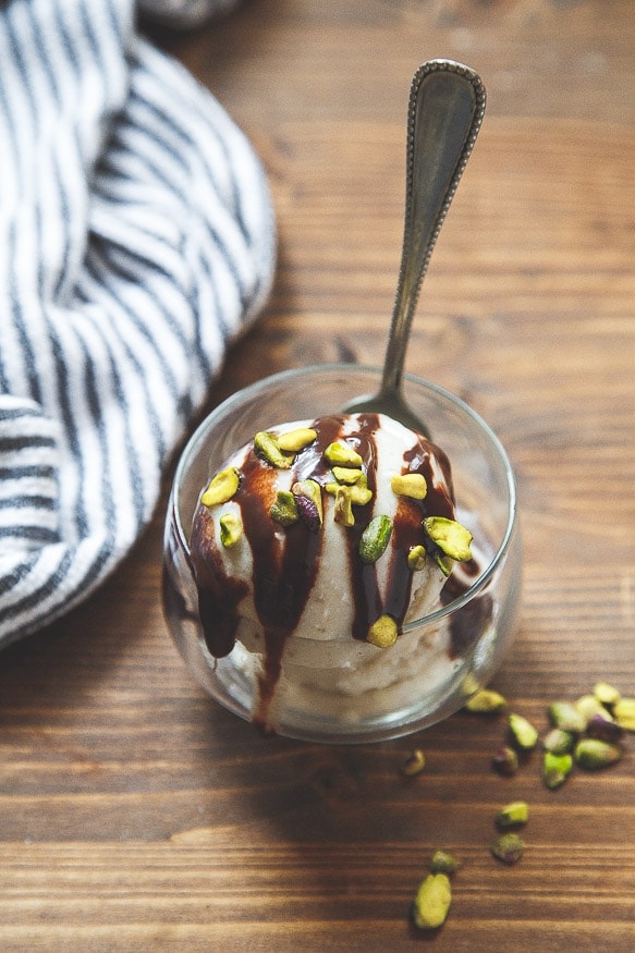 A glass bowl of banana ice cream topped with chocolate sauce and chopped pistachios, with a spoon in the bowl.
