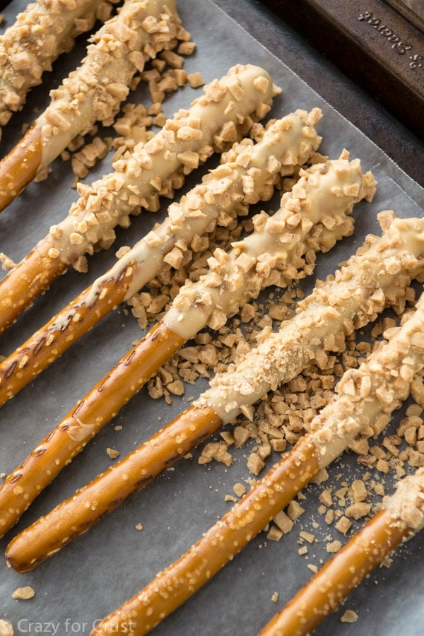 A row of pretzels dipped half way in a tan colored dipped, coated in toffee pieces. 