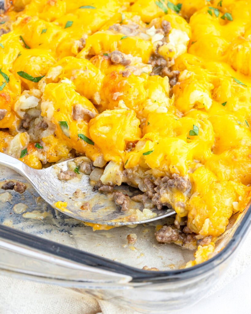 A baking pan of tater tot casserole topped with melted cheese and fresh parsley, with a scoop of the casserole in a spoon.