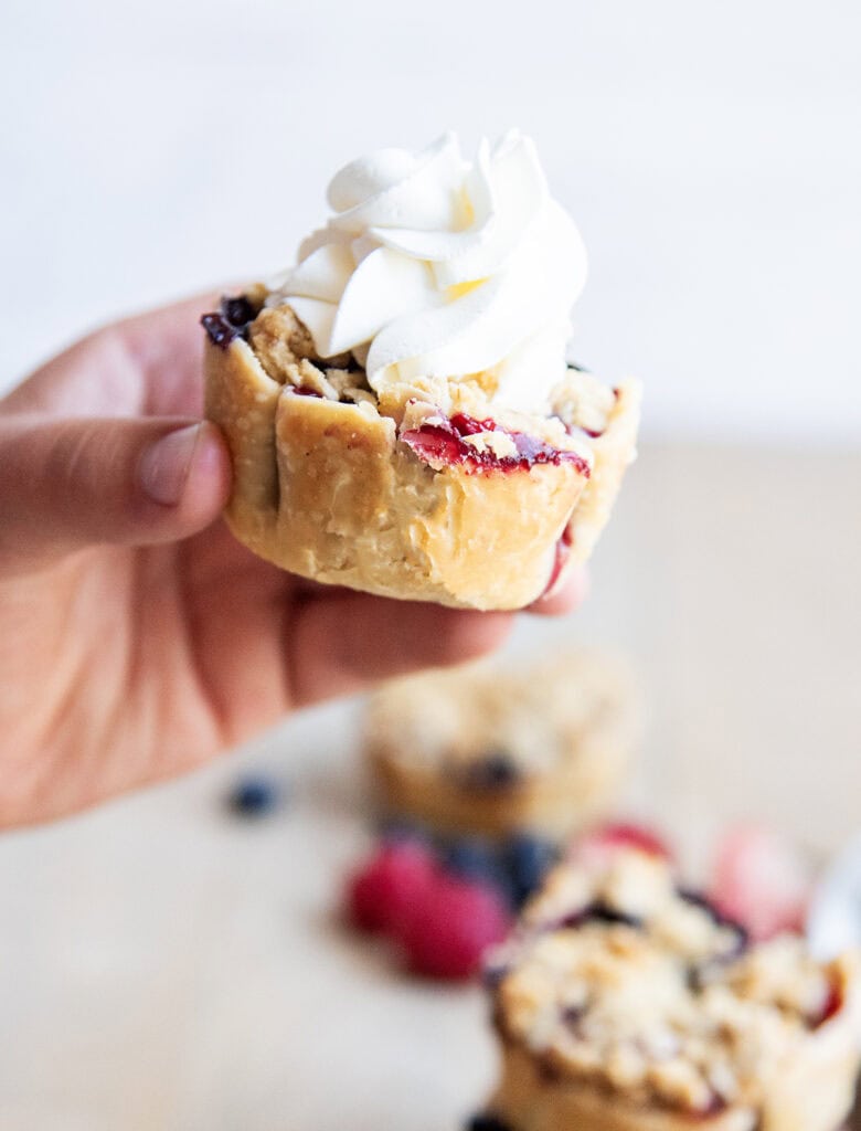 A hand holding a mini berry pie topped with whipped cream.