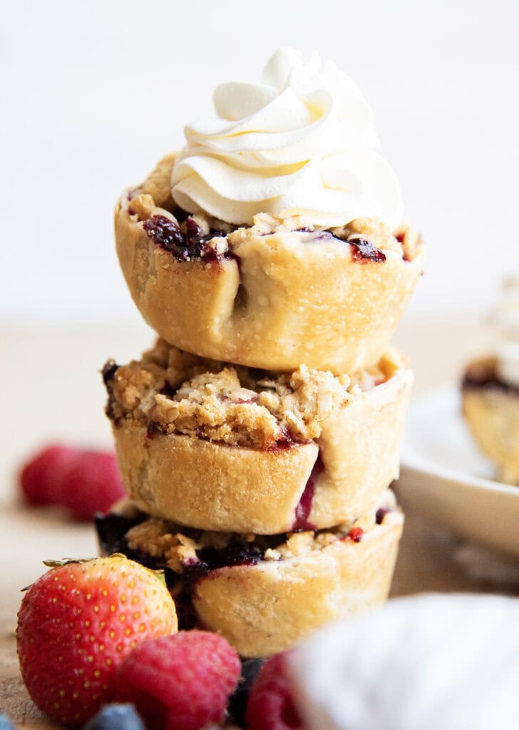 A stack of three crumble topped berry pies, with the top pie loaded with whipped cream.
