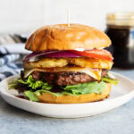 A teriyaki cheeseburger on a plate with pineapple, tomato, and lettuce on it.
