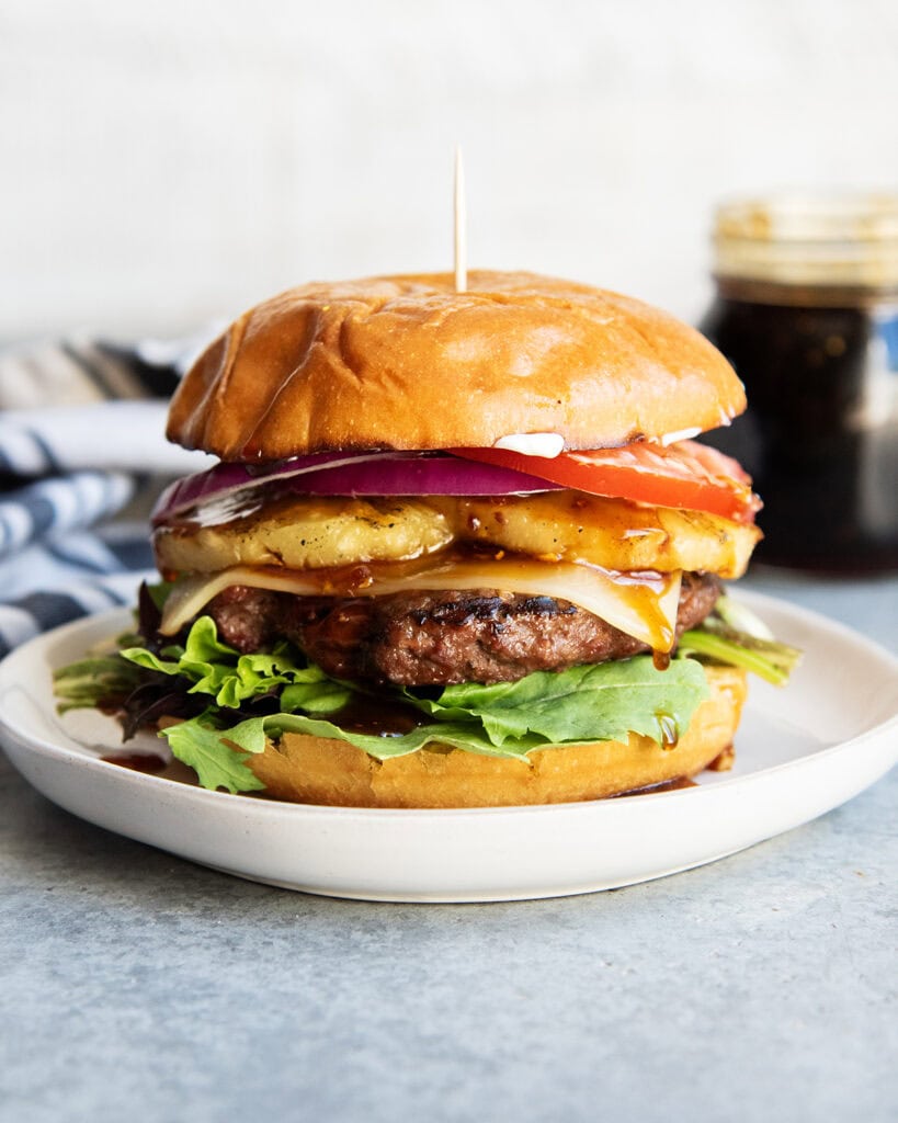 A teriyaki cheeseburger on a plate with pineapple, tomato, and lettuce on it.