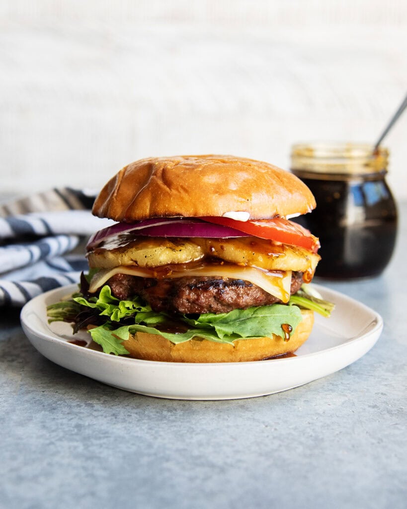 A teriyaki cheeseburger on a plate with pineapple, tomato, and lettuce on it.