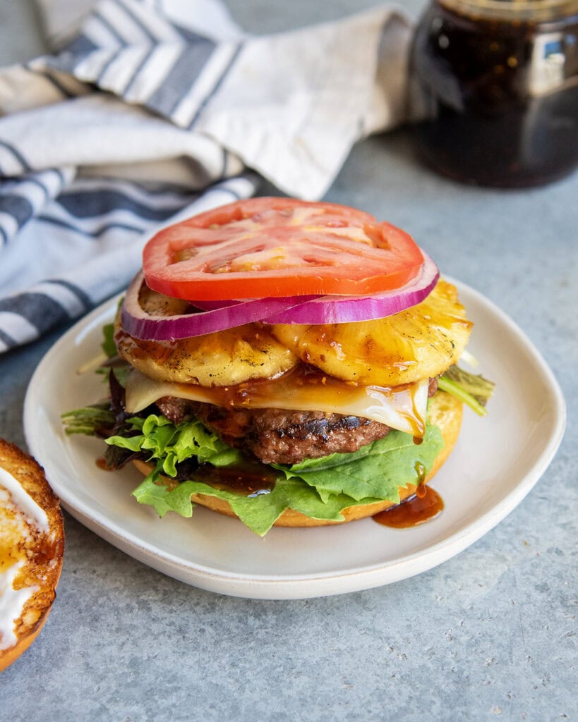 A teriyaki cheeseburger on a plate with pineapple, tomato, and lettuce on it.