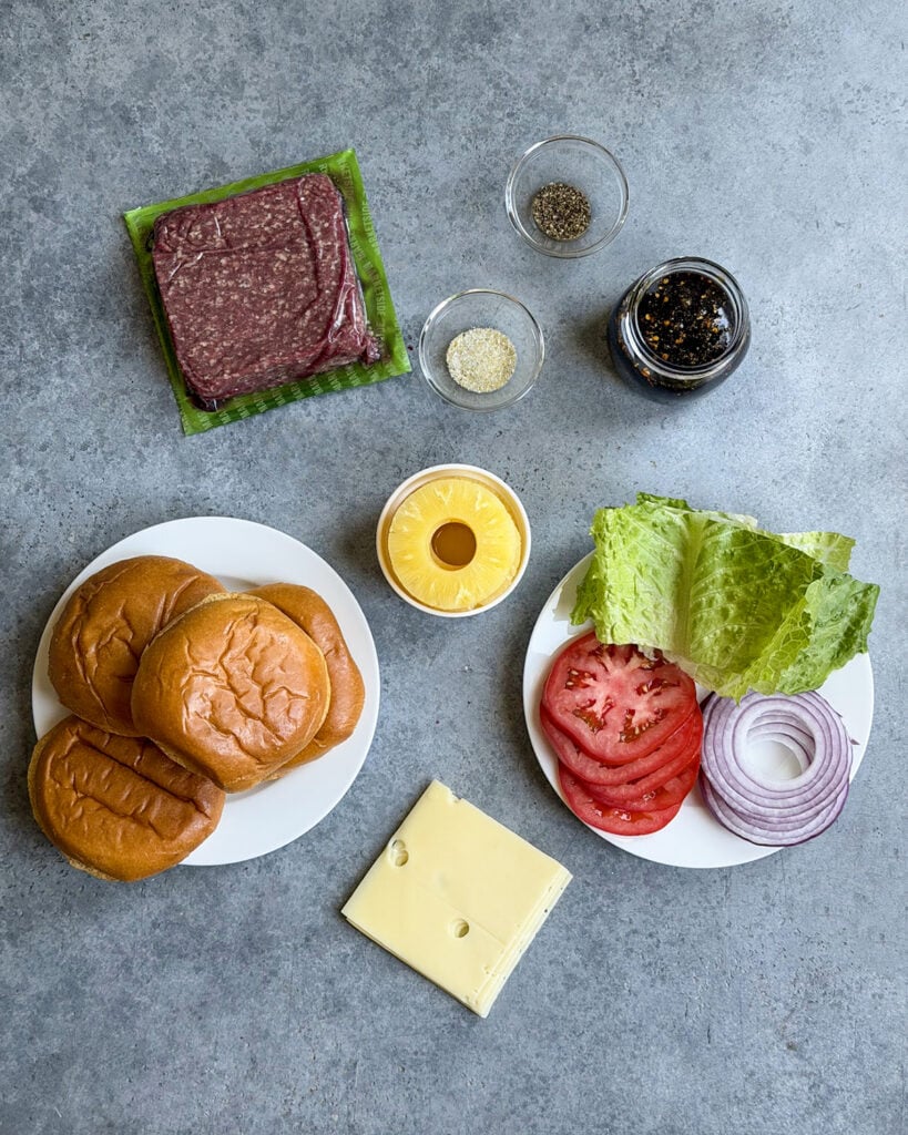 The ingredients needed to make Teriyaki Cheeseburgers, like ground beef, teriyaki sauce, pineapple slices, burger buns, and cheese.