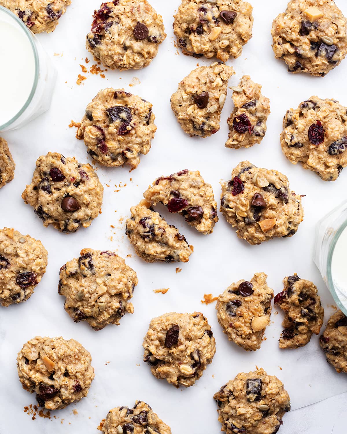 An overhead photo of cookies on a white background, the cookies are arranged randomly, some are split in half, they are full of oats, dried cranberries, and chocolate.