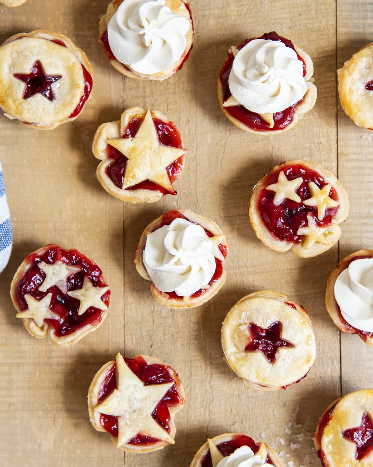 An overhead photo of mini cherry pies topped with star shaped crust pieces. A few of them have whipped cream piped on top.