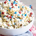 White Chocolate covered Popcorn with red white and blue m&ms and sprinkles in a white bowl