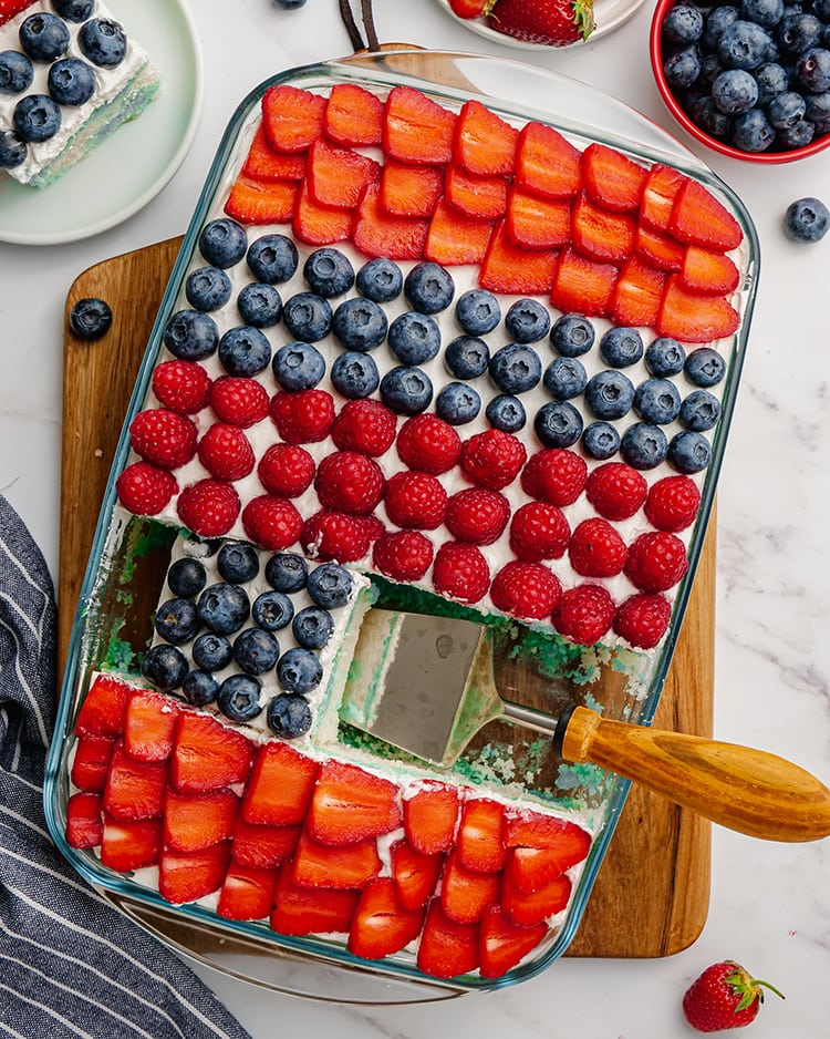 A cake in a pan with stripes of strawberries, blueberries and raspberries with a slice removed.