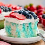 A white cake poked with blue jello and topped with whipped cream and strawberries and blueberries
