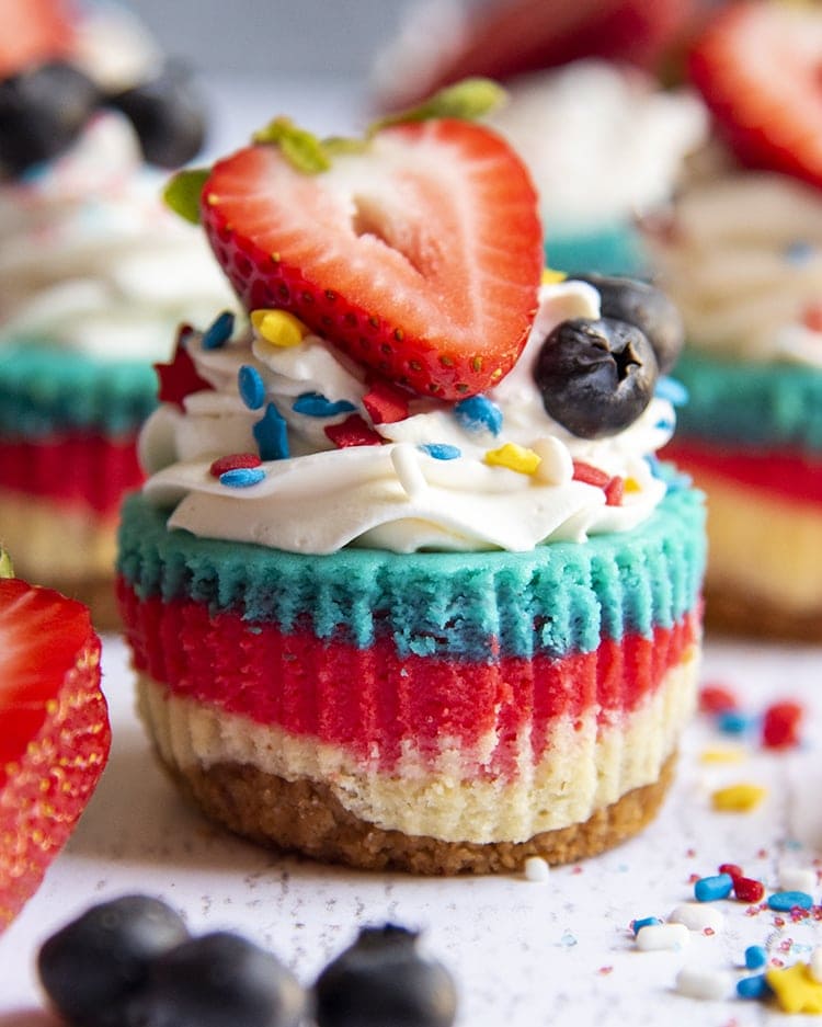 A mini cheesecake with layers of white, red, and blue topped with whipped cream and a fresh strawberry.