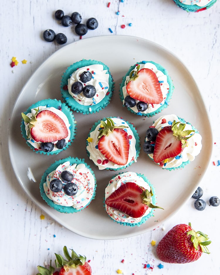 A plate of mini cheesecakes shot from above, showing their blue color topped with whipped cream, and fresh blueberries and strawberries.