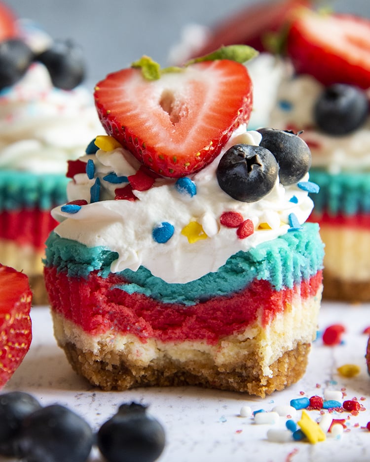 A layered red white and blue mini cheesecake with whipped cream, and a strawberry and 2 blueberries on top, with a bite taken out of it.