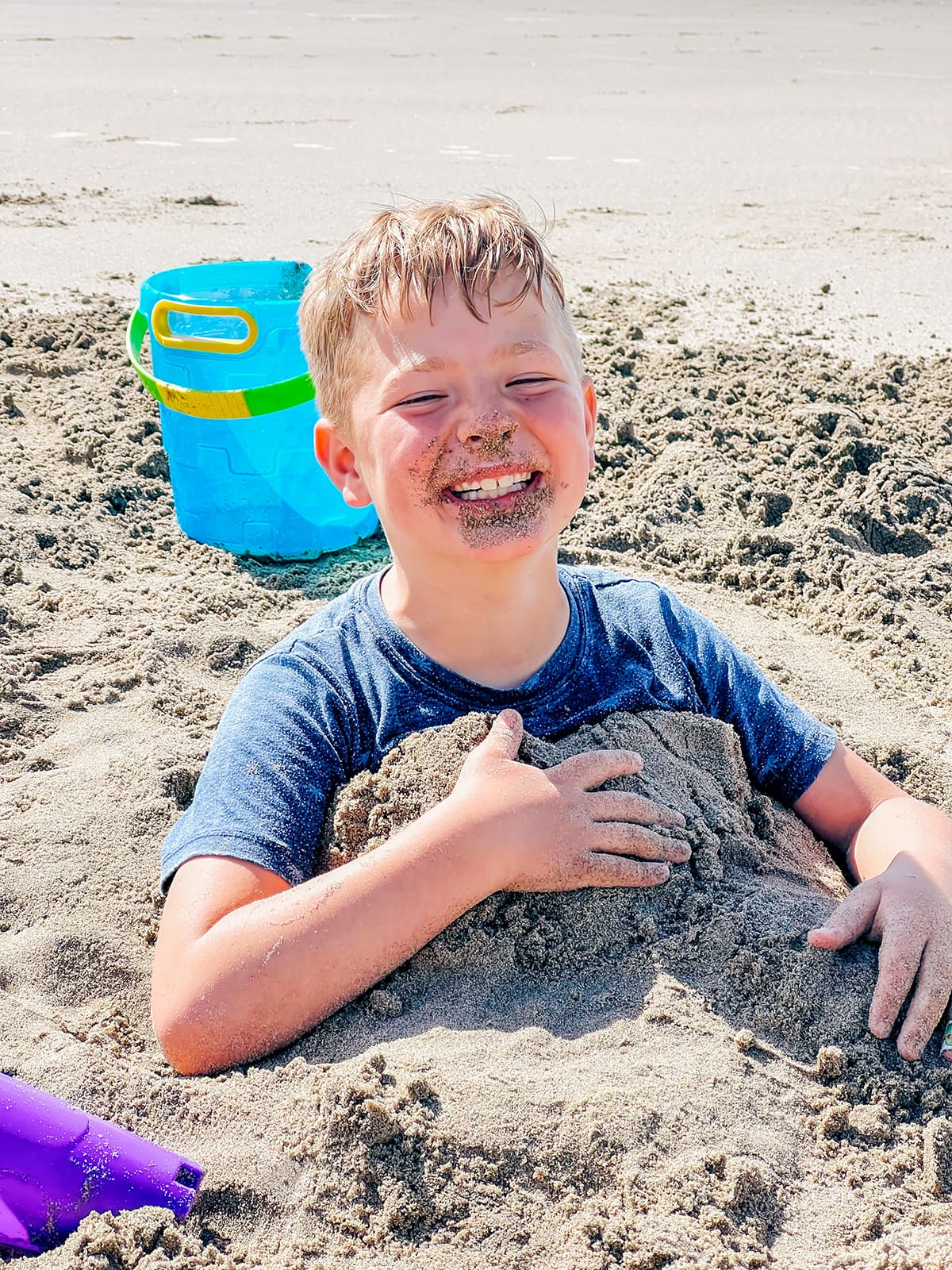 A boy buried in the sand, with sand on his face.
