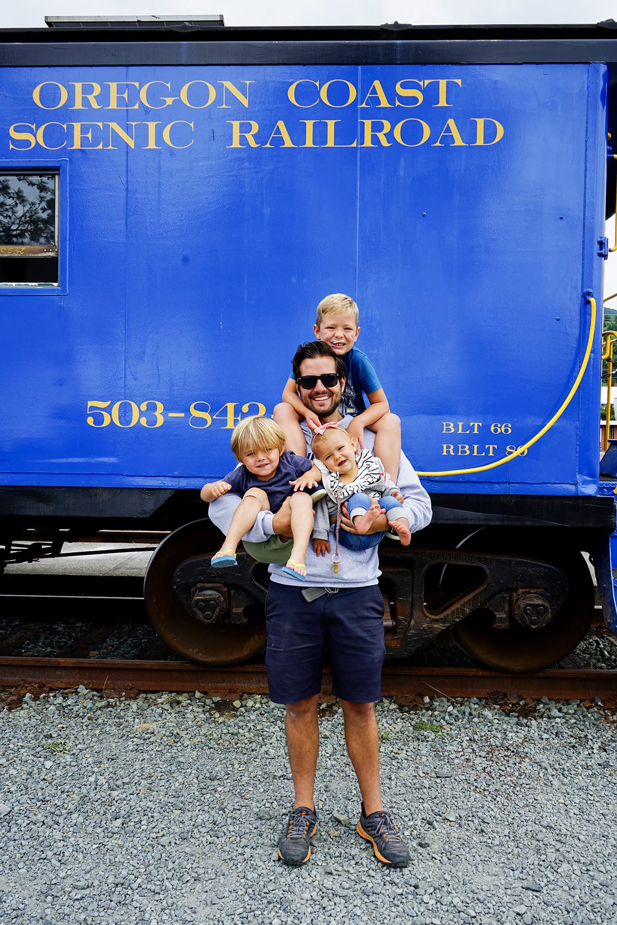 A man holding three children in front of a blue train. 