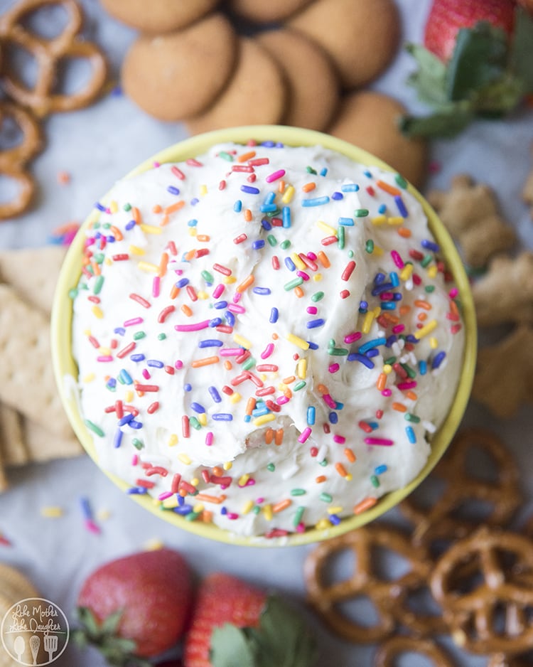 Funfetti cake dip is displayed in a bowl with white sprinkles on top and snacks surrounding the bowl