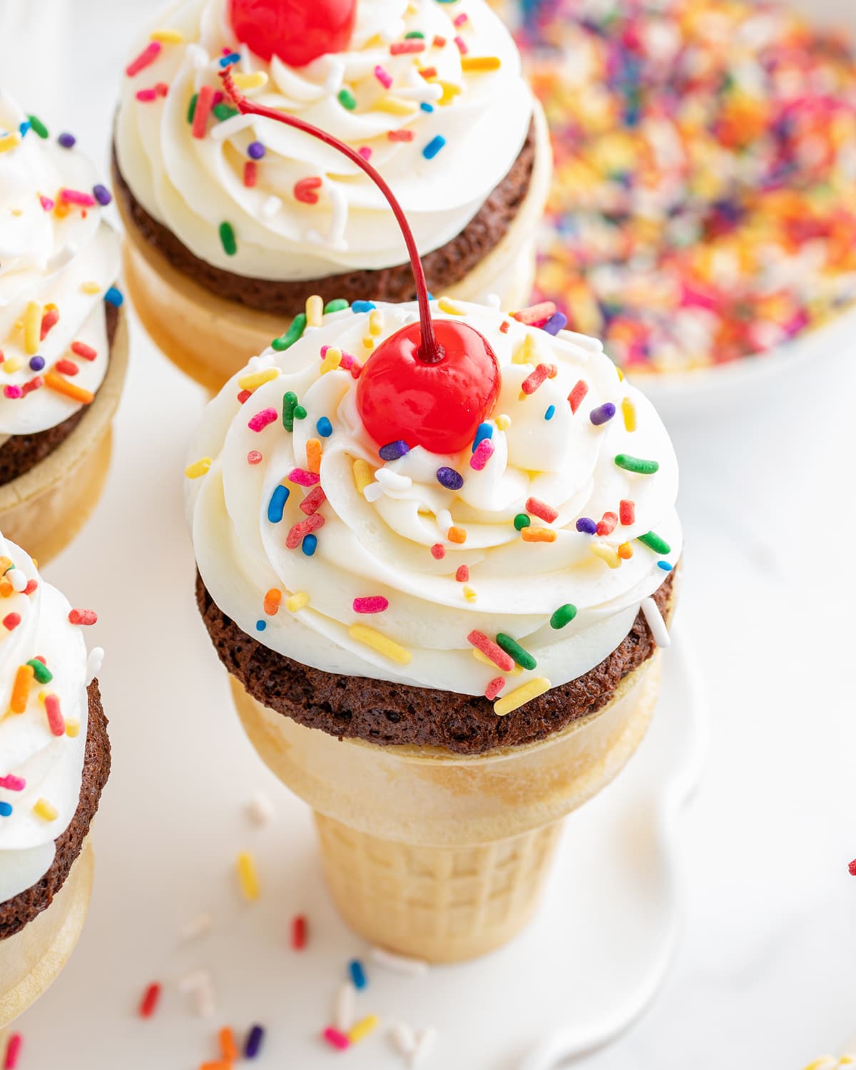 An ice cream cone cupcake topped with a swirl of vanilla frosting and a cherry on top.