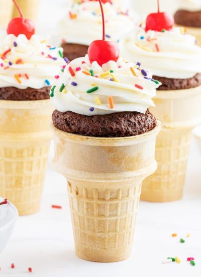 A cupcake in an ice cream cone topped with frosting, sprinkles, and a maraschino cherry.