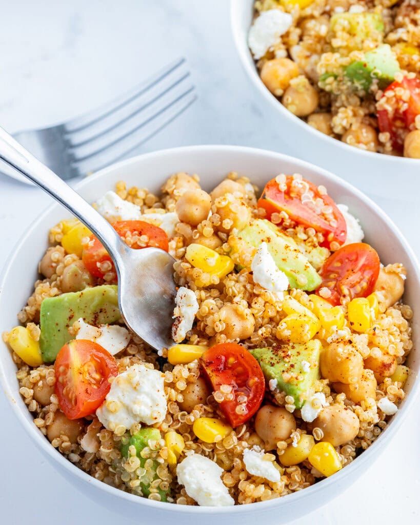 A bowl of a quinoa, chick pea, and corn salad also with tomatoes, avocado, and cotija cheese with a spoon in the bowl.