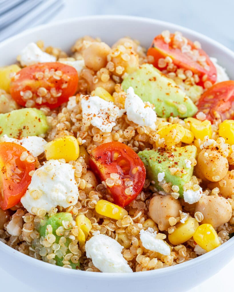A bowl of quinoa and corn salad topped with tomatoes, avocado, and cotija cheese.
