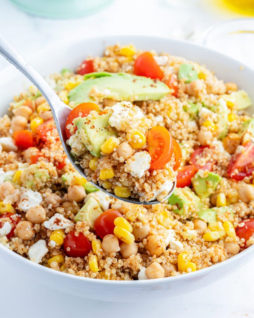 A bowl of a quinoa, chick pea, and corn salad also with tomatoes, avocado, and cotija cheese with a spoon of it above the bowl.