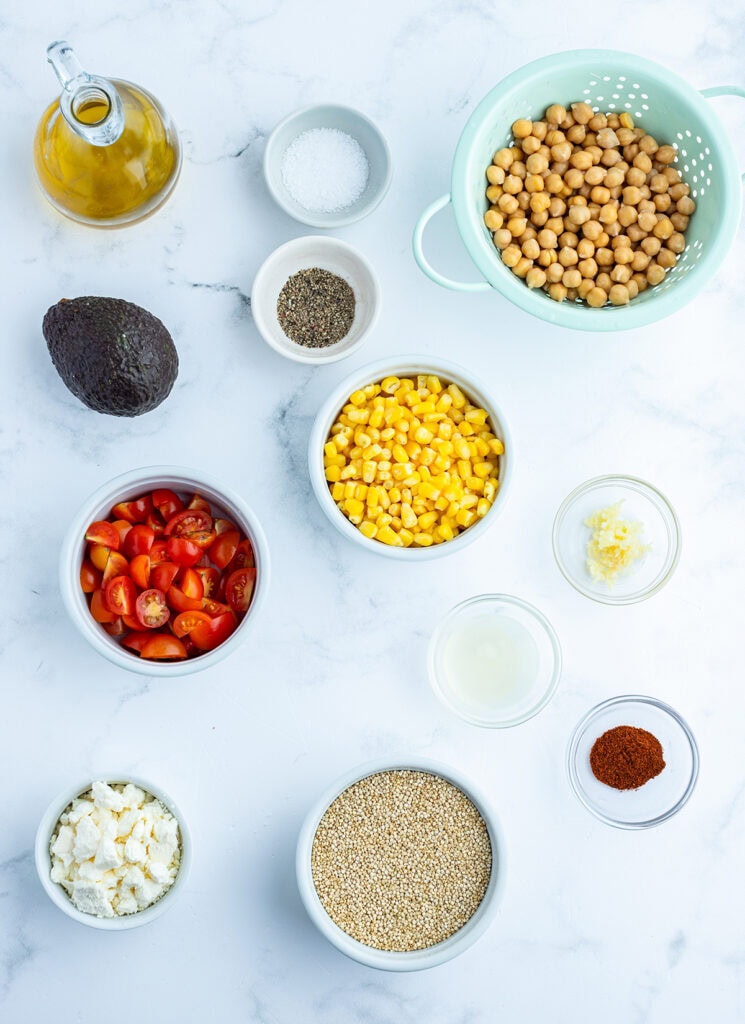 Bowls of the ingredients needed to make corn and quinoa salad, with cherry tomatoes, an avocado, chickpeas, and more. 