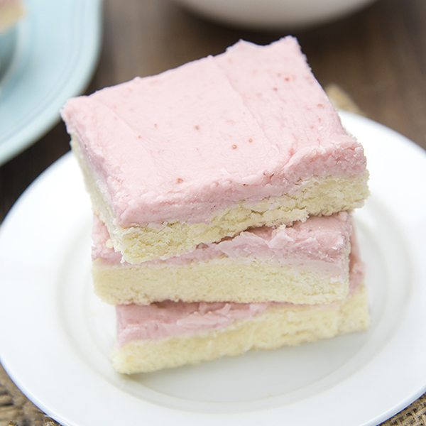 Angled view of strawberry frosted sugar cookie bars stacked on a white plate.