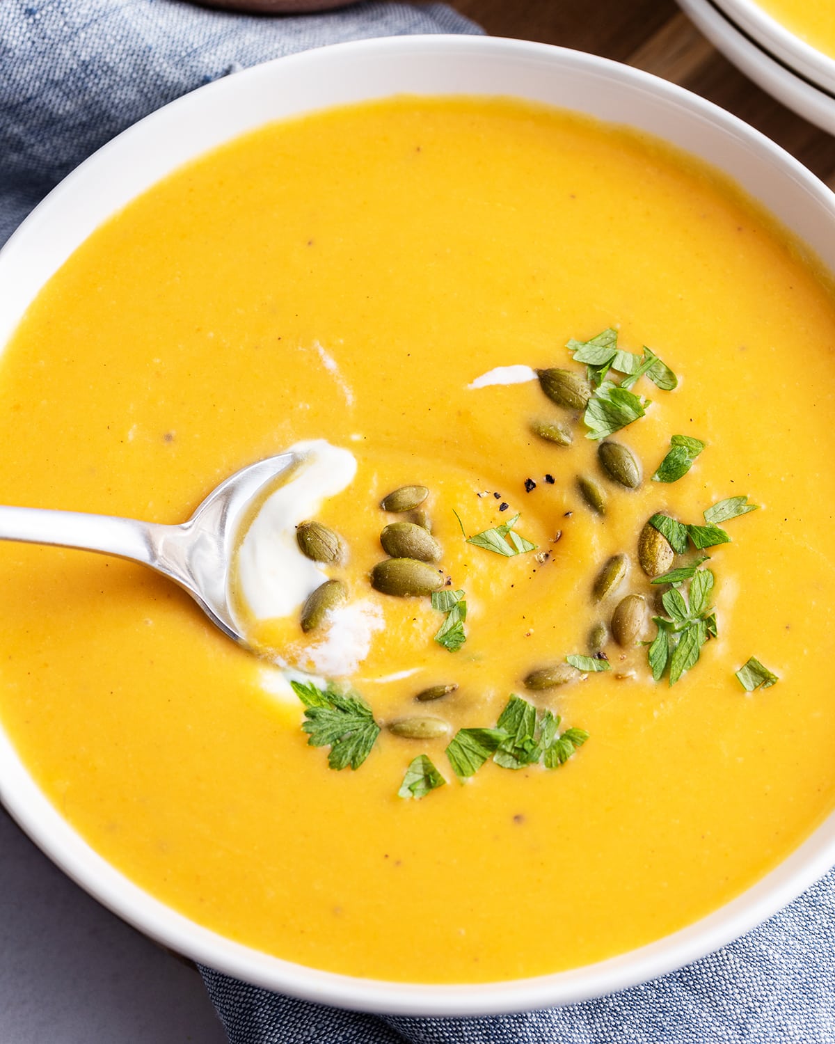 A close up of a bowl of autumn squash soup topped with pepitas and fresh parsley with a spoon in it.