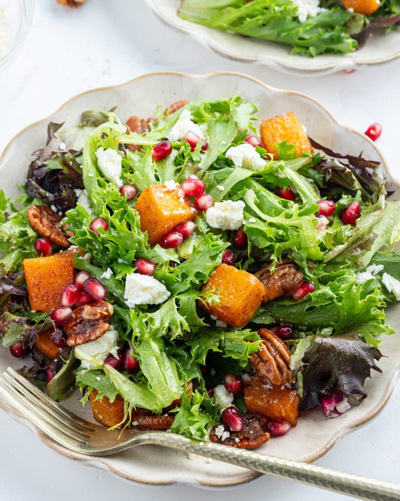 A plate of salad topped with butternut squash pieces, feta, pomegranate arils, and candied pecans with a fork on the plate.