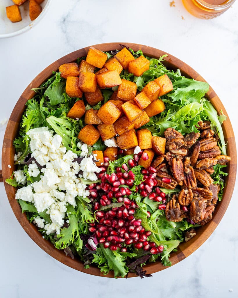 A bowl of salad topped with roasted butternut squash, candied pecans, pomegranate seeds, and feta.