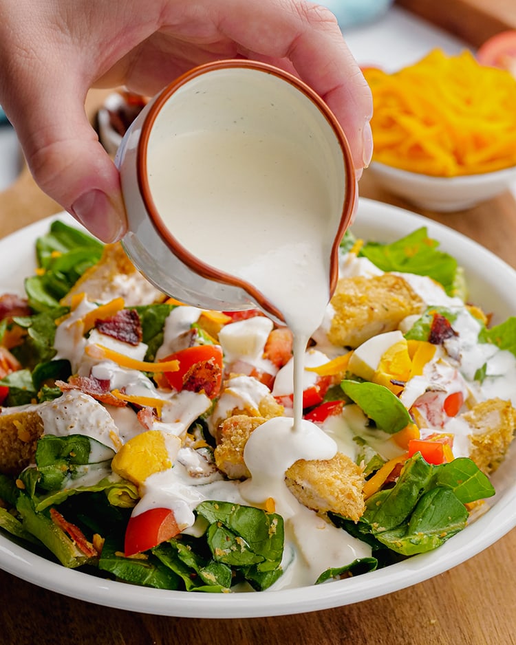 Ranch dressing being poured out of a small cup onto a salad with crispy chicken and diced tomatoes.