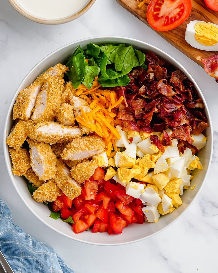 A bowl of ingredients for a cobb salad, crispy chicken, cheese, lettuce, bacon, diced boiled eggs, and diced tomatoes.