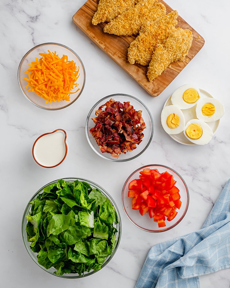 The ingredients in a crispy chicken cobb salad in different bowls before putting it together. A bowl of cheddar cheese, bacon, eggs, tomatoes, lettuce, and ranch dressing, and crispy chicken strips on a cutting board.