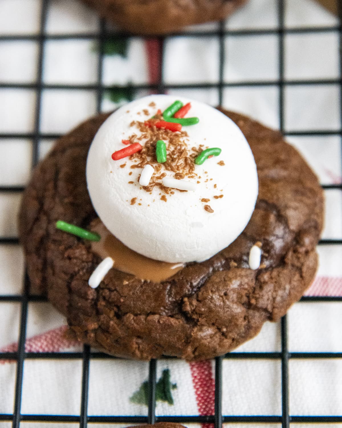 A close up of a hot chocolate cookie topped with a marshmallow and sprinkles on a wire rack.