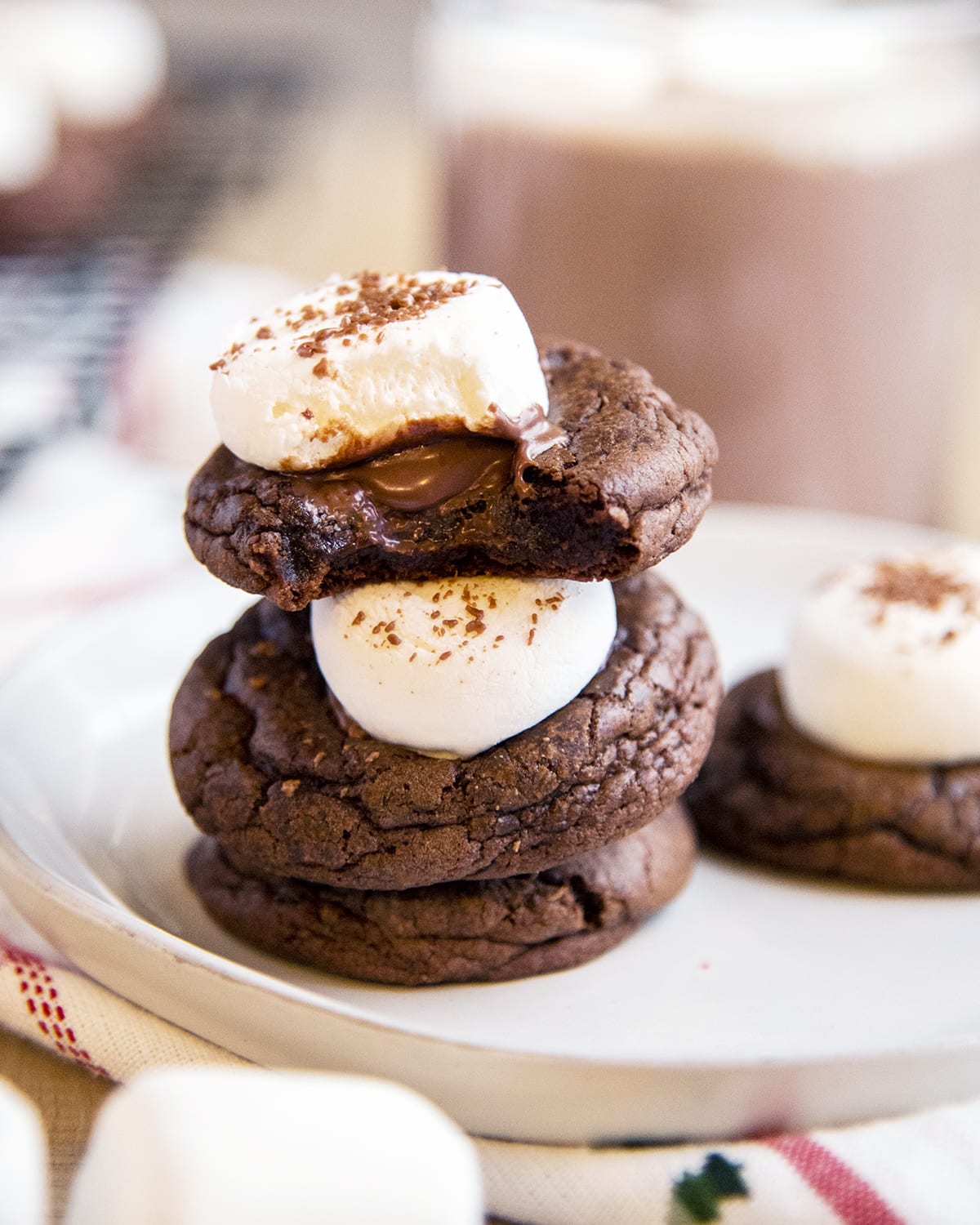 A stack of three hot chocolate cookies topped with gooey marshmallows, and the top cookie is bit in half showing the melted chocolate underneath.
