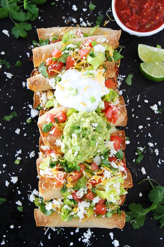 A bunch of taquitos lined up next to one another topped with lettuce, pico, avocado, sour cream, and cilantro. 