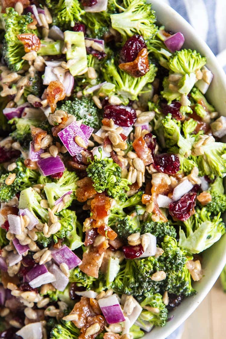 A close up of broccoli salad, with chopped broccoli, red onion, dried cranberries, sunflower seeds, and bacon, with a light creamy dressing.