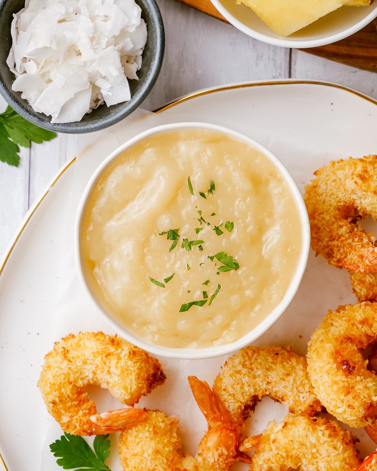 A small bowl of coconut shrimp sauce, that is a light yellow color, the bowl is on a plate surrounded by coconut shrimp.